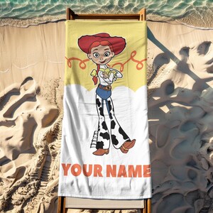 May include: A rectangular beach towel featuring a cartoon image of Jessie from Toy Story. The towel has a yellow top with Jessie's image and a white bottom with the text "YOUR NAME" in orange. The towel is on a wooden beach chair.