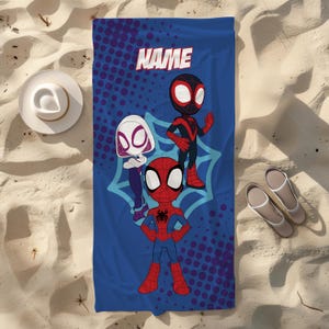 May include: Blue beach towel with cartoon Spider-Man characters. The towel has the word "NAME" at the top. The characters include Spider-Man, Spider-Gwen, and Miles Morales. The towel is on a sandy surface with a hat and sandals.