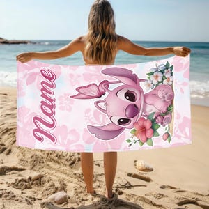 Pode incluir: Uma toalha de praia rosa com uma personagem de desenho animado, uma borboleta e detalhes florais. A toalha tem a palavra "Name" em it&aacute;lico. O fundo mostra uma cena de praia com areia e o mar.