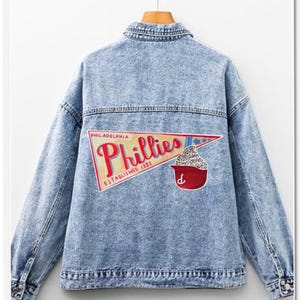 Peut inclure: Veste en jean bleu clair avec un motif au dos. Le motif comprend le mot "Phillies" en écriture rouge, un bol rouge et le texte "Philadelphia Established 1883". La veste est suspendue à un cintre en bois.