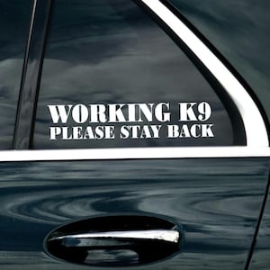 May include: A white vinyl decal with the text "WORKING K9 PLEASE STAY BACK" on a dark blue car window.