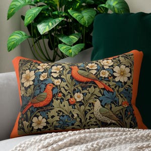 May include: Rectangular cushion with an orange border. It features a floral and bird design in blue, green, orange, and white. The cushion is on a grey sofa with a white blanket. Decorative home decor.