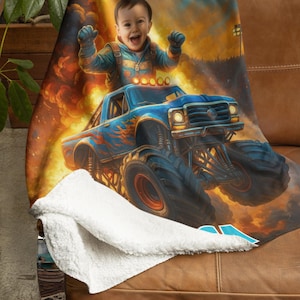 May include: A plush throw blanket featuring a vibrant graphic print of a smiling child with arms raised, riding in a blue monster truck with orange flame decals. The background shows a fiery explosion. The blanket has a white sherpa fleece lining.