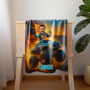 May include: A rectangular blanket featuring a graphic print of a child with arms raised, sitting atop a blue monster truck with orange flame decals. The background shows an explosion with fire and smoke. The name LUCA is printed in blue letters.
