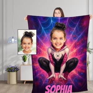 May include: A rectangular fleece blanket featuring a custom illustration of a child with a high bun hairstyle, depicted as a superhero in a white, pink, and black suit. The background shows a glowing spiderweb over a nebula, with the name SOPHIA.