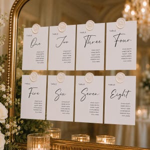 May include: Eight white rectangular wedding seating chart cards arranged in two rows on a gold-framed mirror. Each card features a table number in black script, a list of names, and a circular cream-colored wax seal with a floral design at the top.