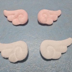 May include: Four felt angel wings, two pink and two white, are arranged on a light blue surface. The wings are about 2 inches long and 1 inch wide.
