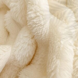May include: Close-up of a cream-colored, plush blanket. The soft, fluffy texture is visible, with long, dense fibers creating a warm and inviting appearance. The blanket's folds and shadows add depth to the image.