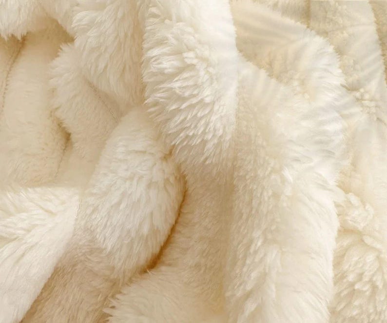 May include: Close-up of a cream-colored, plush blanket. The soft, fluffy texture is visible, with long, dense fibers creating a warm and inviting appearance. The blanket's folds and shadows add depth to the image.