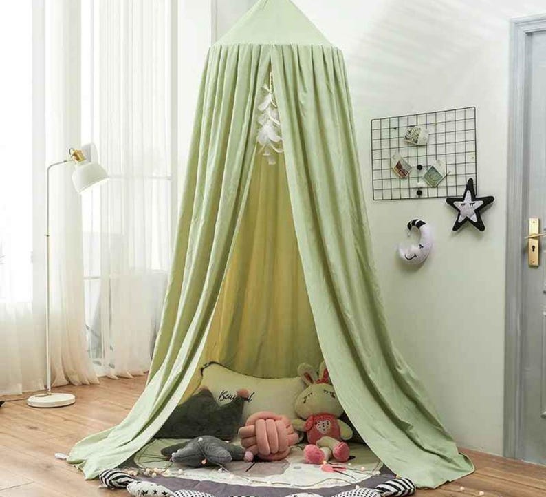 May include: A light green canopy bed with draped fabric, creating a cozy space. Inside, there are plush toys and pillows on a patterned rug. The canopy is suspended from the ceiling, offering a whimsical touch to a child's room.