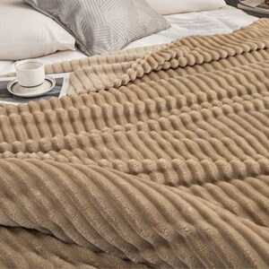 May include: A tan, textured blanket with a ribbed design covers a bed. A white teacup and saucer sit on a magazine. Pillows are visible in the background.