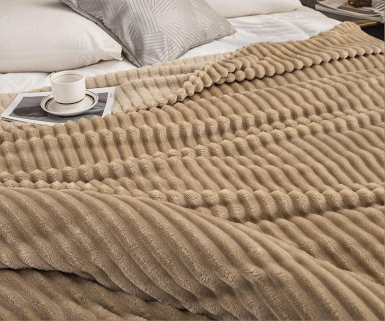 May include: A tan, textured blanket with a ribbed design covers a bed. A white teacup and saucer sit on a magazine. Pillows are visible in the background.