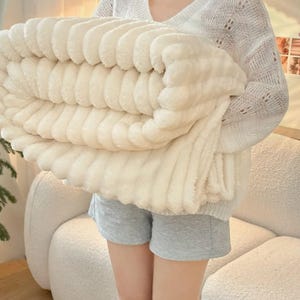 May include: A folded, cream-colored plush blanket with a ribbed texture. The blanket is held by a person wearing a white sweater and gray shorts. The blanket appears soft and cozy, suitable for home decor.