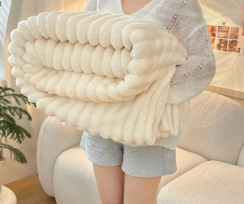 May include: A folded, cream-colored plush blanket with a ribbed texture. The blanket is held by a person wearing a white sweater and gray shorts. The blanket appears soft and cozy, suitable for home decor.
