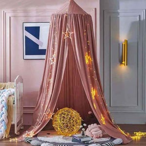 May include: A pink canopy bed with star-shaped lights hanging from the top. A gold sphere and a pink pillow are on the floor. A white crib with a braided bumper is on the left.