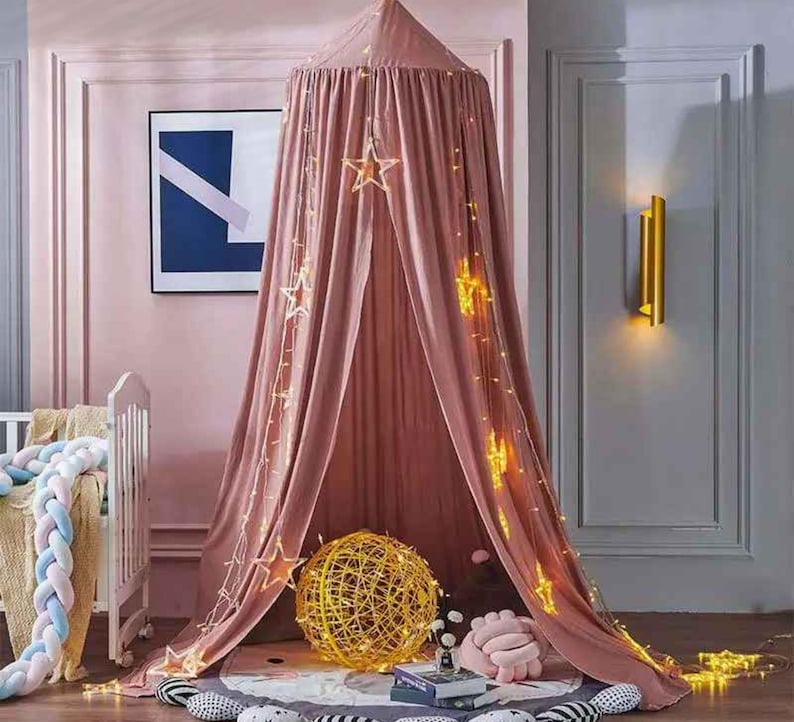 May include: A pink canopy bed with star-shaped lights hanging from the top. A gold sphere and a pink pillow are on the floor. A white crib with a braided bumper is on the left.