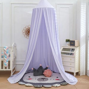 May include: A light purple canopy hangs from the ceiling, draped over a round rug with a lion face design. A gray crown pillow and a pink knot pillow rest on the rug. A white crib and a small dresser are also in the room.
