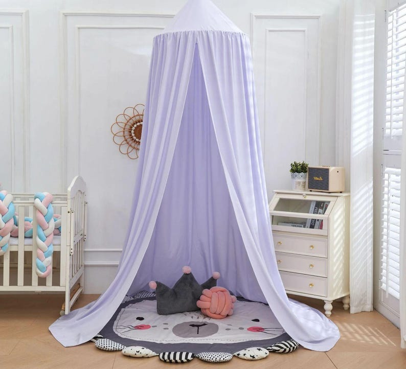 May include: A light purple canopy hangs from the ceiling, draped over a round rug with a lion face design. A gray crown pillow and a pink knot pillow rest on the rug. A white crib and a small dresser are also in the room.