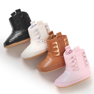 May include: Four infant boots arranged in a diagonal row, featuring black, white, tan, and light pink colours. Each boot has a smooth finish, a pull tab at the top, and decorative stitching on the shaft and toe area with a flat, flexible sole.