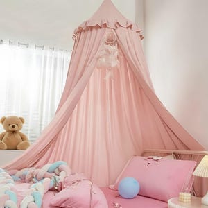 May include: A soft pink fabric bed canopy featuring a ruffled top edge and a hanging dreamcatcher with white feathers. The canopy drapes over a bed with a pink pillow, a braided pastel bumper, a blue balloon, and a small white lamp nearby.