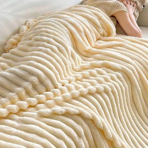 May include: A cream-coloured, plush blanket with a ribbed design. The blanket is draped over a person, creating a cosy and inviting scene. The soft texture and neutral colour suggest comfort and warmth, perfect for relaxation.