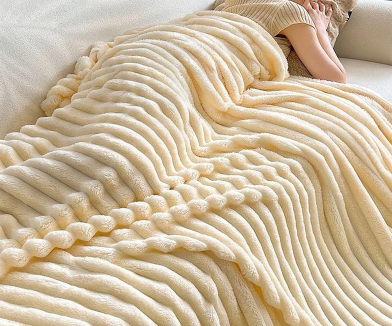 May include: A cream-colored, plush blanket with a ribbed design. The blanket is draped over a person, creating a cozy and inviting scene. The soft texture and neutral color suggest comfort and warmth, perfect for relaxation.