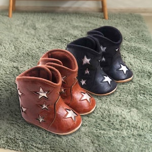 May include: Two pairs of baby cowboy boots, one brown and one black, with star cutouts and star detailing. The boots are made from a soft material and are designed for infants. They are arranged on a green rug.
