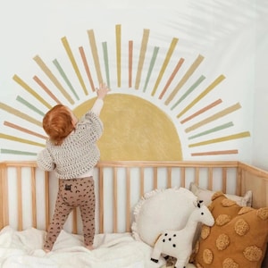 Puede incluir: Un niño con un suéter de crochet y pantalones estampados alcanza un diseño de sol pintado en una pared blanca. La habitación cuenta con una cuna de madera con almohadas y un peluche.
