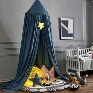 May include: A navy blue canopy with a yellow star decoration, creating a cozy play area. Inside, there are star and crown-shaped pillows, a teddy bear, and a decorative light. A white crib and framed artwork are in the background.