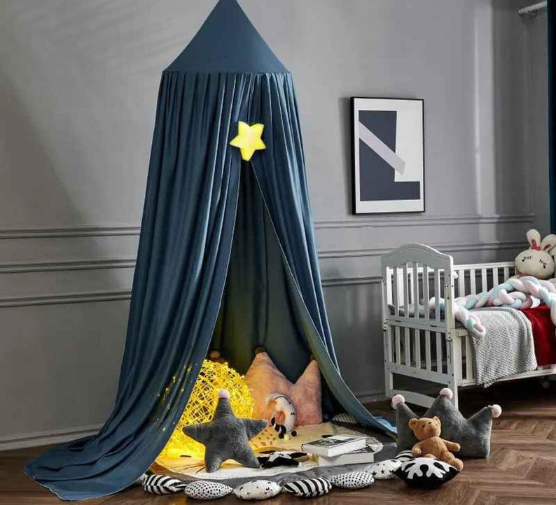 May include: A navy blue canopy with a yellow star decoration, creating a cozy play area. Inside, there are star and crown-shaped pillows, a teddy bear, and a decorative light. A white crib and framed artwork are in the background.
