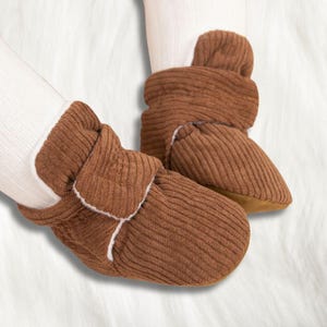 Könnte beinhalten: Ein Paar braune Baby-Booties aus Cord mit weißem Besatz. Die Booties haben ein weiches, strukturiertes Aussehen und sind so konzipiert, dass sie die Füße des Babys warm halten. Die Sohlen sind hellbraun.