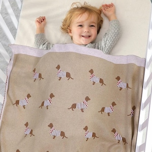 Può includere: Una coperta beige per bambini con un motivo ripetuto di bassotti marroni con camicie a righe. La coperta ha un bordo rosa chiaro ed è in una culla bianca. Un bambino sorride nella culla.