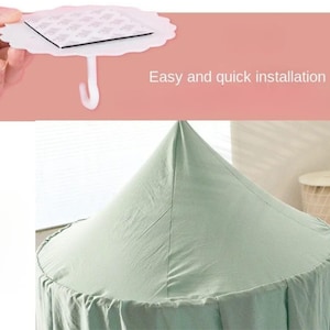May include: A white, scalloped-edge adhesive hook with a square adhesive pad and a curved hook, held by a hand. The text "Easy and quick installation" is displayed. Below is a light green canopy.