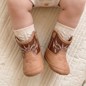 May include: Baby cowboy boots in tan and brown with decorative stitching. The boots feature a classic western design and are paired with cream cable knit socks. These infant boots are designed for little feet.