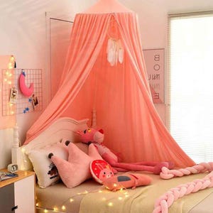 May include: A coral pink canopy bed with a dream catcher and decorative pillows. The bed is adorned with string lights and a pink braided bumper. A pink panther stuffed animal is on the bed.