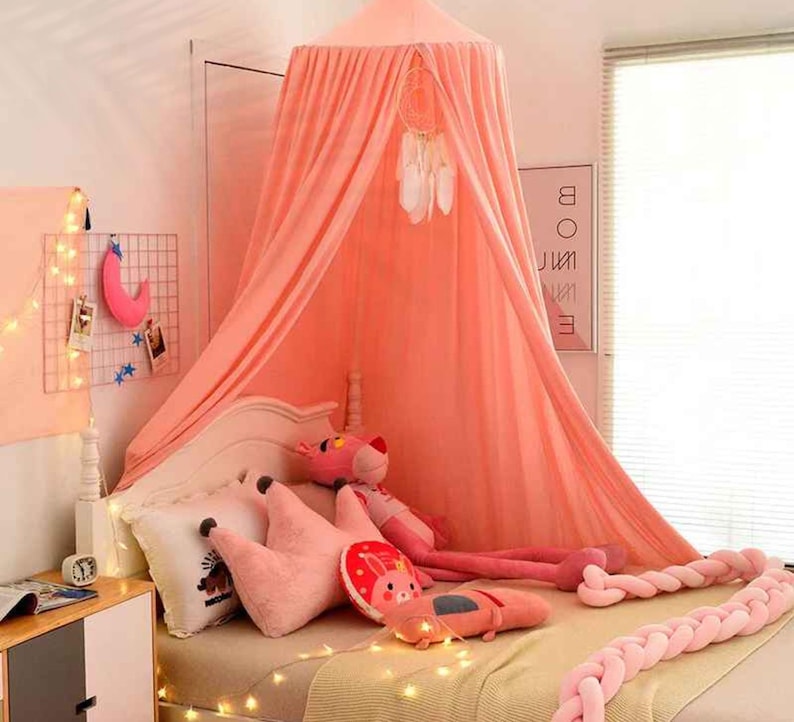 May include: A coral pink canopy bed with a dream catcher and decorative pillows. The bed is adorned with string lights and a pink braided bumper. A pink panther stuffed animal is on the bed.