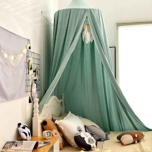 May include: A mint green canopy bed with draped fabric, creating a cozy space. The bed is adorned with decorative pillows and stuffed animals. A small wooden table holds books and a clock. A wall grid displays photos and decor.