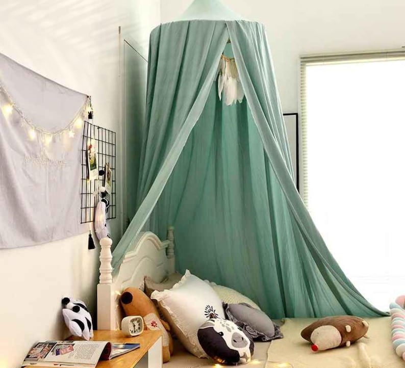 May include: A mint green canopy bed with draped fabric, creating a cozy space. The bed is adorned with decorative pillows and stuffed animals. A small wooden table holds books and a clock. A wall grid displays photos and decor.