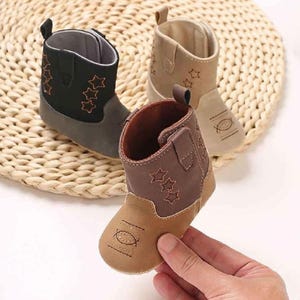 May include: Three infant boots in black, beige, and brown, each featuring a pull tab at the heel, a side opening, and embroidered star patterns on the shaft. The toe area of each boot displays a stitched oval design, and the soles are flat.