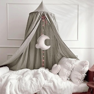 Puede incluir: Una cama con dosel gris con ropa de cama y almohadas blancas. Una almohada blanca en forma de media luna cuelga del dosel. La cama está en una habitación con paredes blancas y un marco de madera roja.