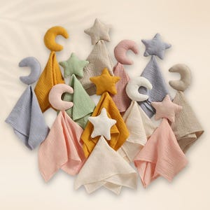 Puede incluir: Una colección de artículos para bebés en colores pastel, que incluye gasas y formas de luna y estrella de peluche. Las gasas son en tonos de amarillo, rosa, verde y azul. Las formas de peluche son de colores a juego.