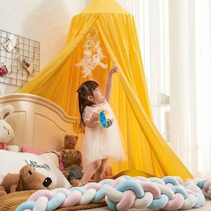 May include: A yellow canopy bed with a dream catcher hanging inside. A young child in a white dress is reaching up towards the dream catcher. A braided bed bumper in pastel colors is at the foot of the bed.
