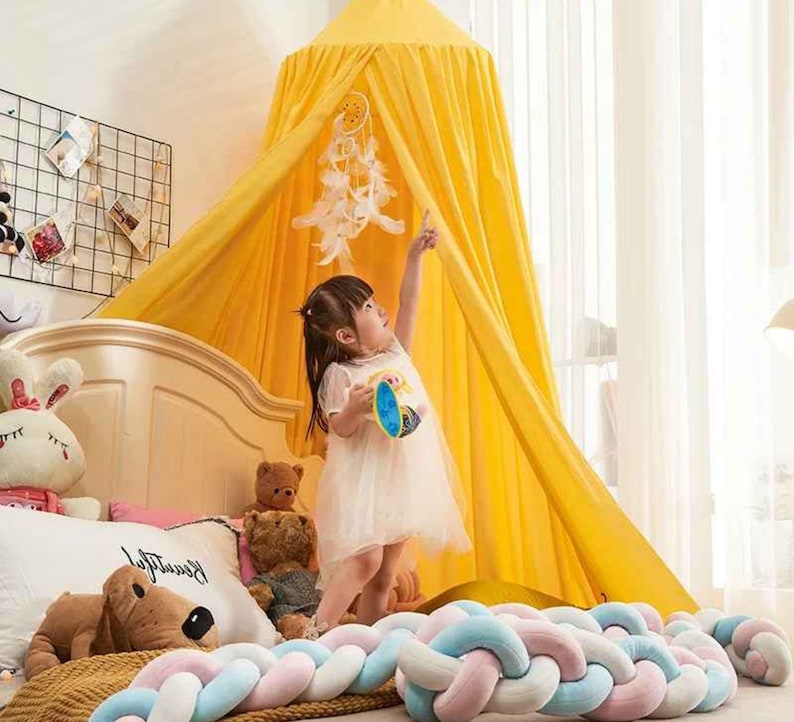 May include: A yellow canopy bed with a dream catcher hanging inside. A young child in a white dress is reaching up towards the dream catcher. A braided bed bumper in pastel colors is at the foot of the bed.