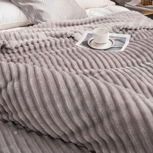 May include: A soft, gray, ribbed blanket covers a bed, with a white pillow and a patterned pillow visible. A white teacup and saucer sit on a newspaper on top of the blanket. The blanket has a plush, textured surface.