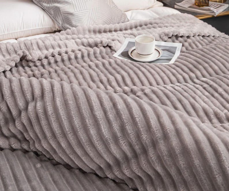 May include: A soft, gray, ribbed blanket covers a bed, with a white pillow and a patterned pillow visible. A white teacup and saucer sit on a newspaper on top of the blanket. The blanket has a plush, textured surface.