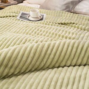 May include: A light green, ribbed blanket covers a bed. A white teacup and saucer sit on a small, rectangular tray on top of the blanket. The blanket has a soft, plush texture and is folded over the bed.