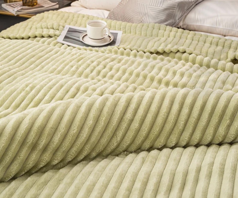May include: A light green, ribbed blanket covers a bed. A white teacup and saucer sit on a small, rectangular tray on top of the blanket. The blanket has a soft, plush texture and is folded over the bed.