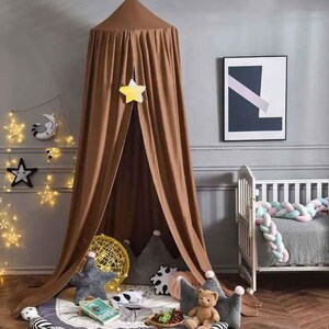 May include: A brown canopy bed tent with a star-shaped decoration hanging from the top. The tent is set up in a nursery with star-shaped string lights, decorative pillows, and a crib. A framed abstract art piece hangs on the wall.