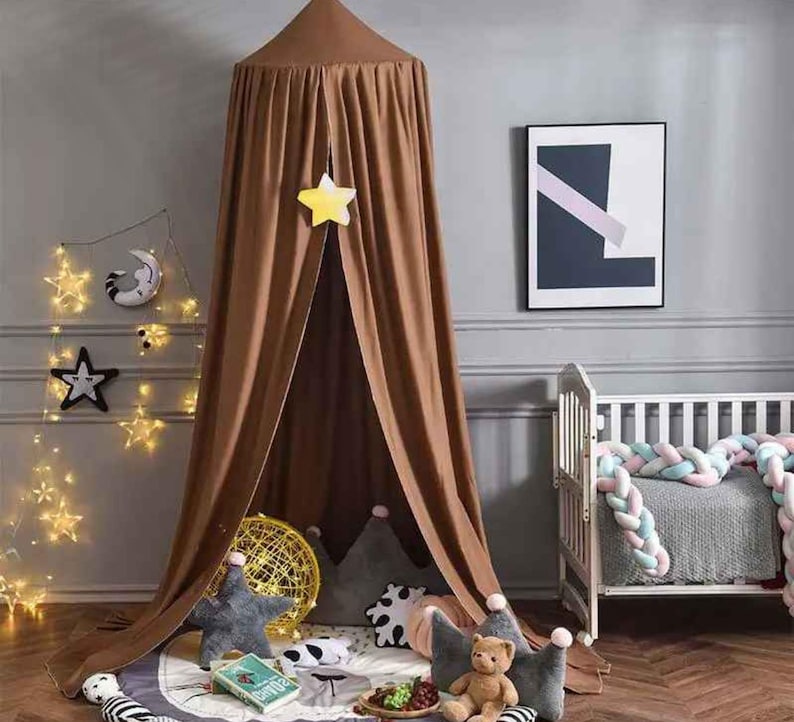 May include: A brown canopy bed tent with a star-shaped decoration hanging from the top. The tent is set up in a nursery with star-shaped string lights, decorative pillows, and a crib. A framed abstract art piece hangs on the wall.