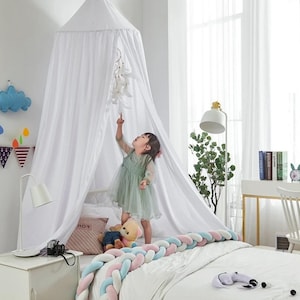 May include: A white canopy bed with a sheer, draped design. A young child in a green dress stands on the bed, reaching towards a hanging mobile. The bed is decorated with a braided bumper in pastel colors.